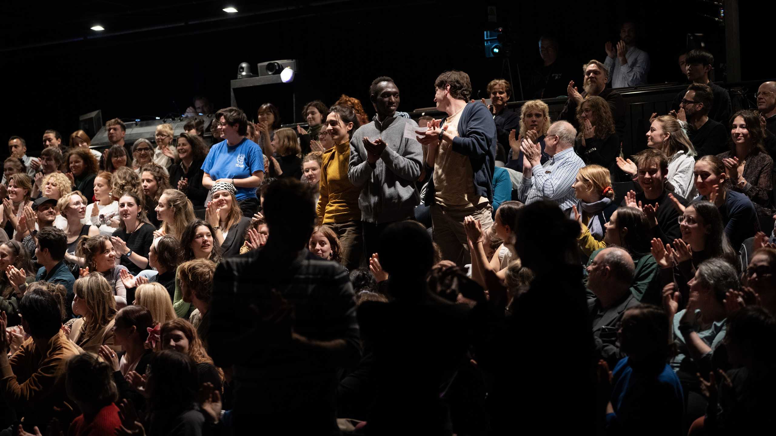 Studierende in einem Theaterraum applaudiert, teils stehend, in einem dunklen Saal