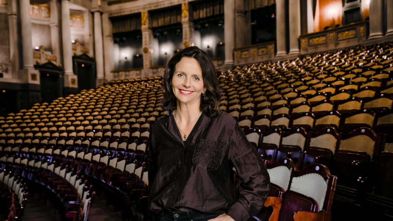 Foto von Amélie Niermeyer mit dunklem Hemd, steht lächelnd im großen, leeren Prinzregententheater, hinter ihr viele Sitzreihen